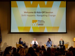 Four women are seated on chairs on a stage. The screen overhead reads: Welcome and Kick-Off Session, Shift Happens: Navigating Change, Women's Leadership Conference 2023. The sign on the podium reads: .