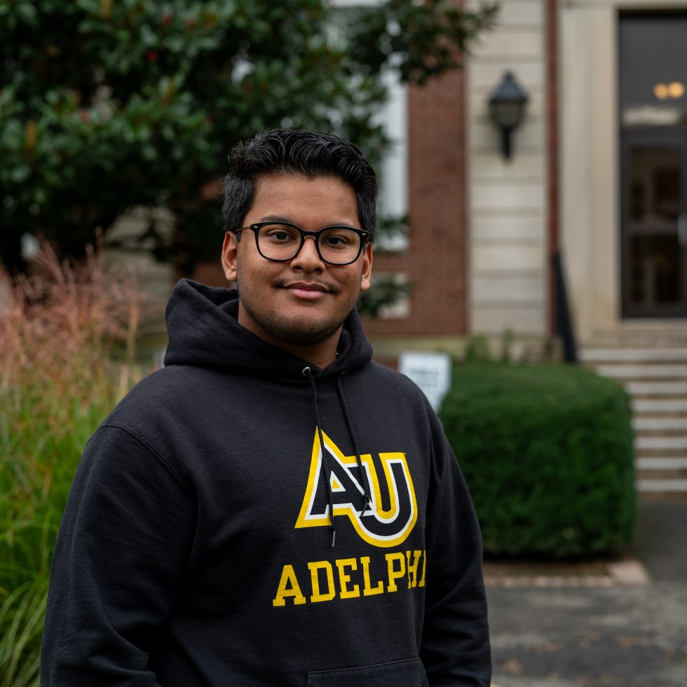 A confident male  student stands in front of Levermore Hall wearing an hoodie.